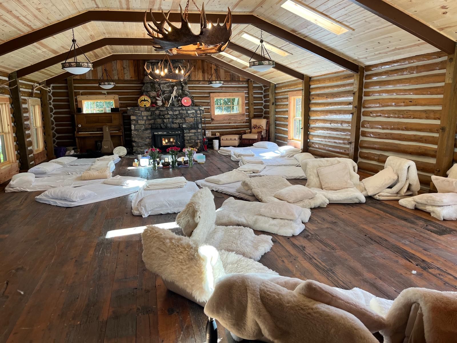 A log cabin room prepared for a group circle, mats and sheepskins arranged around a stone fireplace and flowers