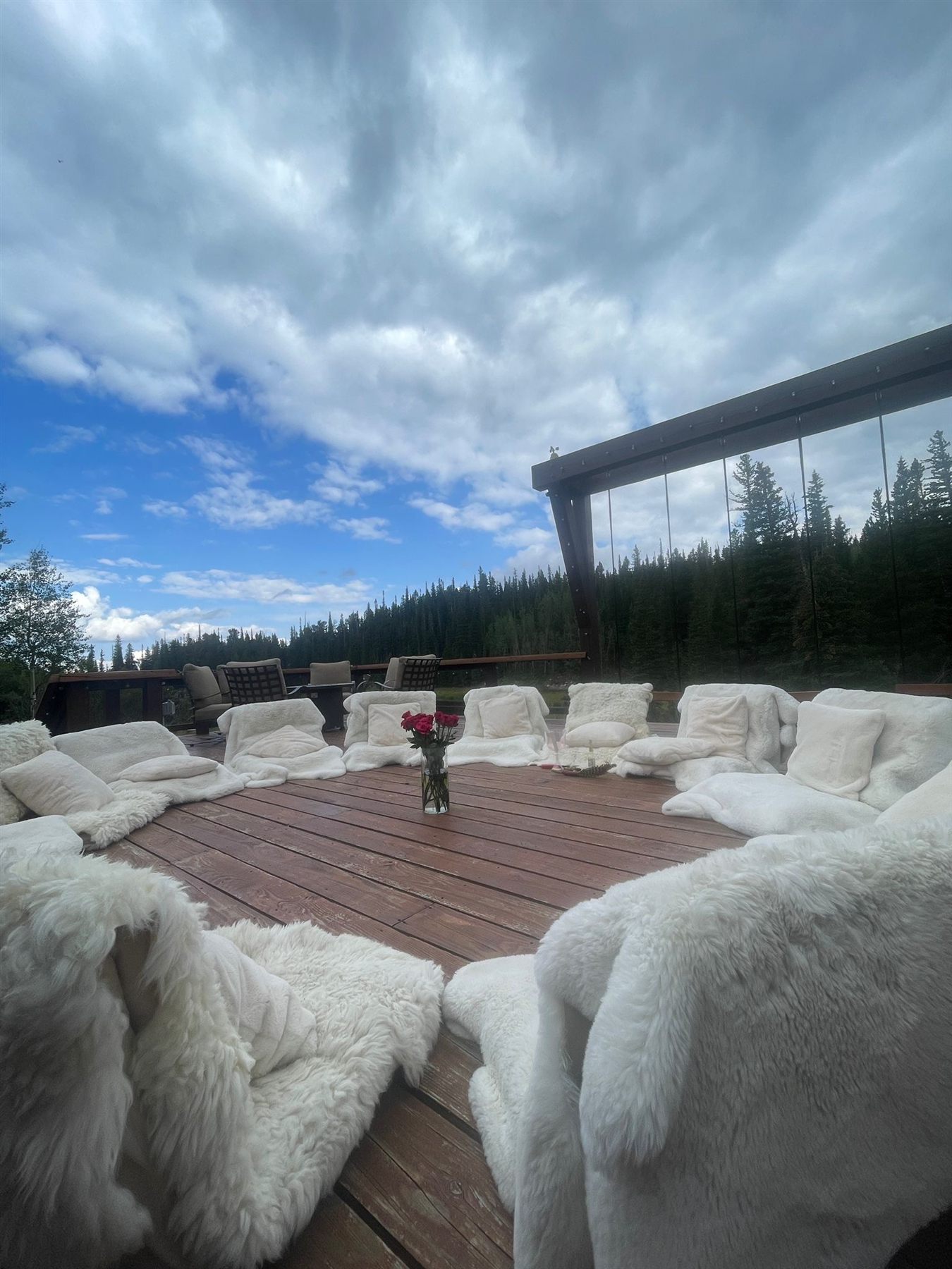 A circle of sheepskin floor seats on a wooden deck under a big mountain sky, a vase of roses in the center