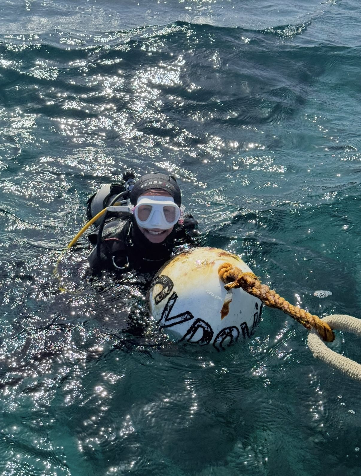 Tracey at the surface in scuba gear, smiling in her mask, one arm over the Gypsy's Jewel mooring buoy