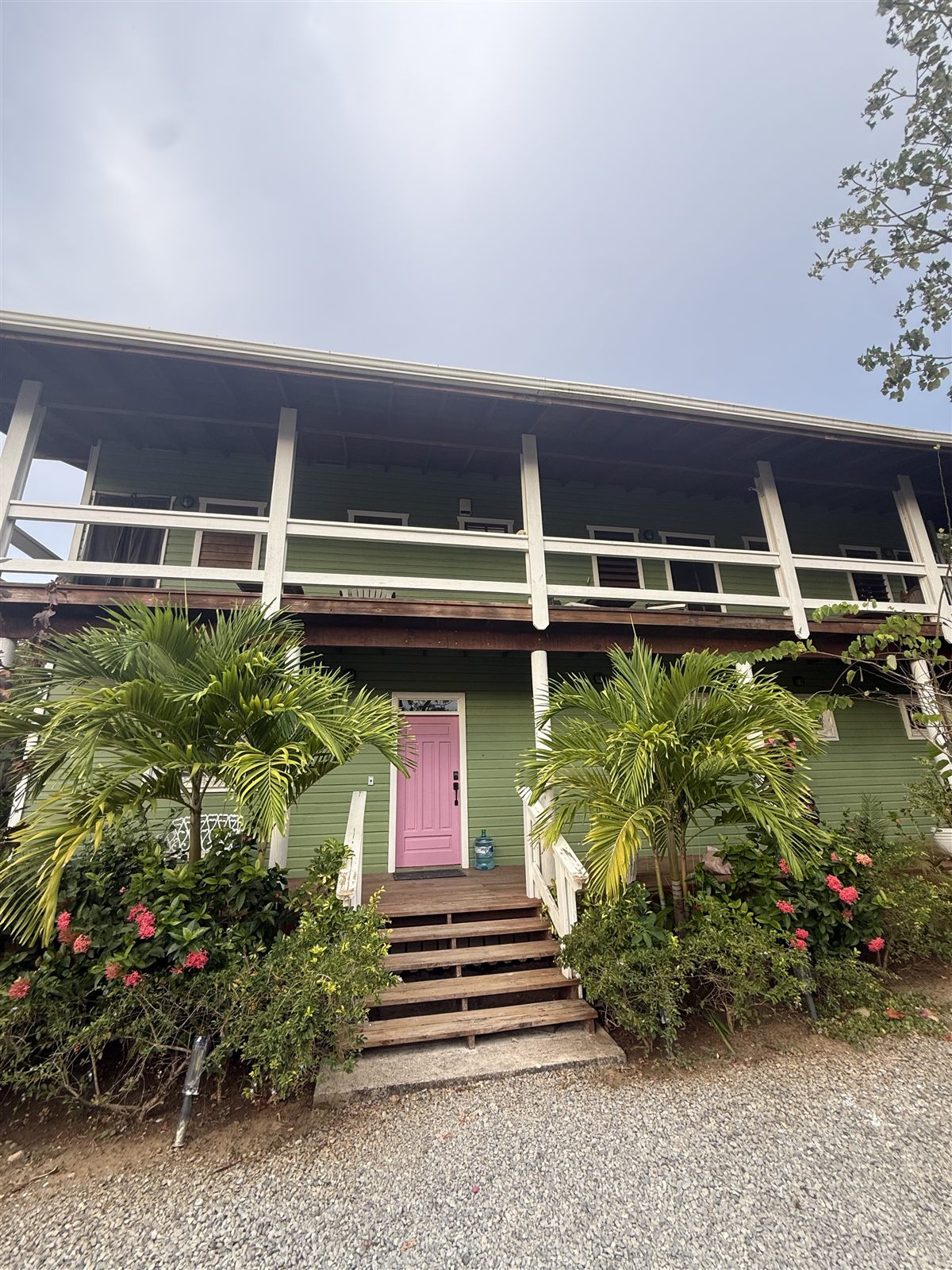 Gypsy's Jewel, the house: a green two story island home with a pink front door, wraparound verandas, and palms