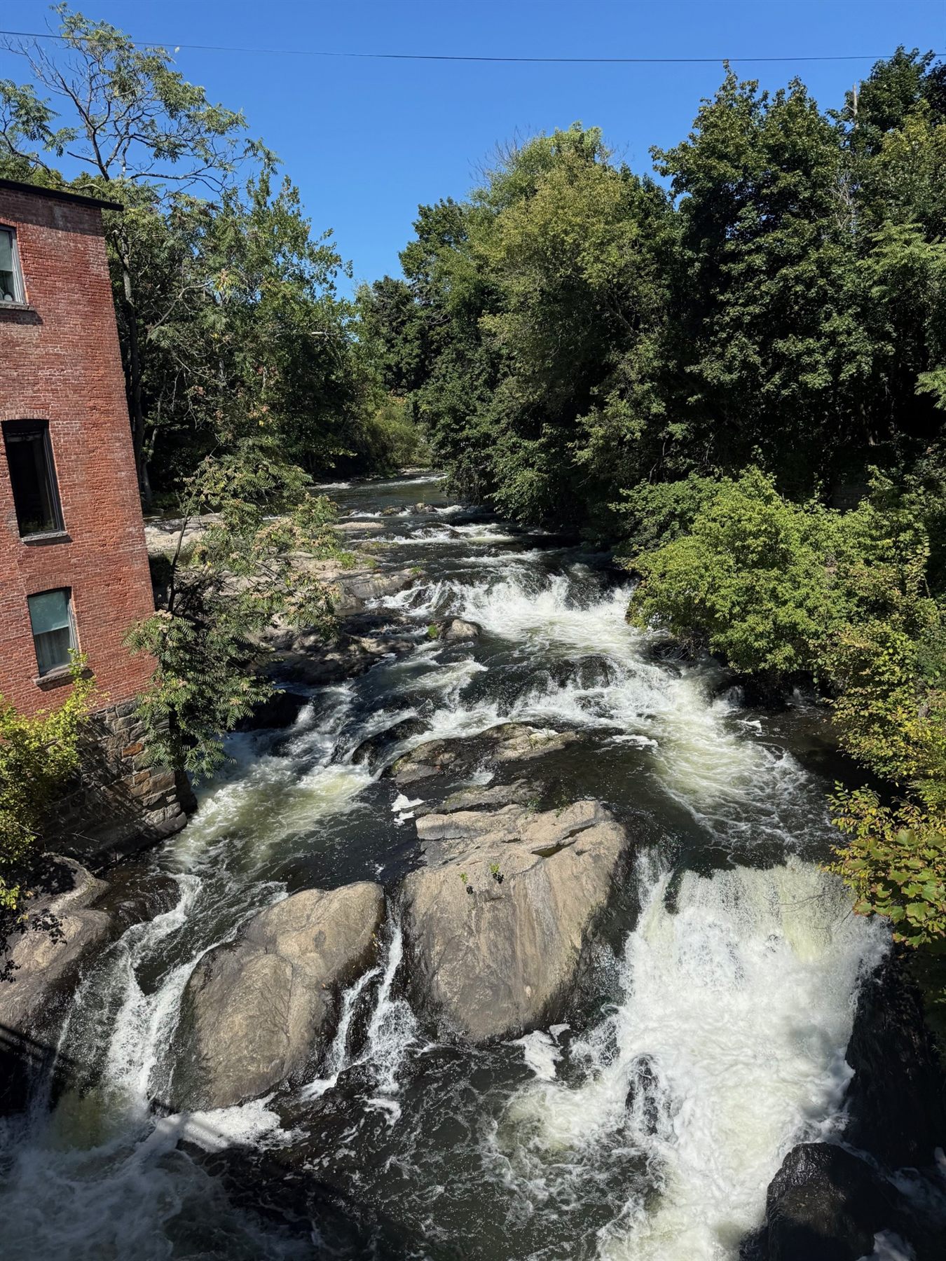 A creek pouring over dark rocks beside an old brick mill, trees on both banks