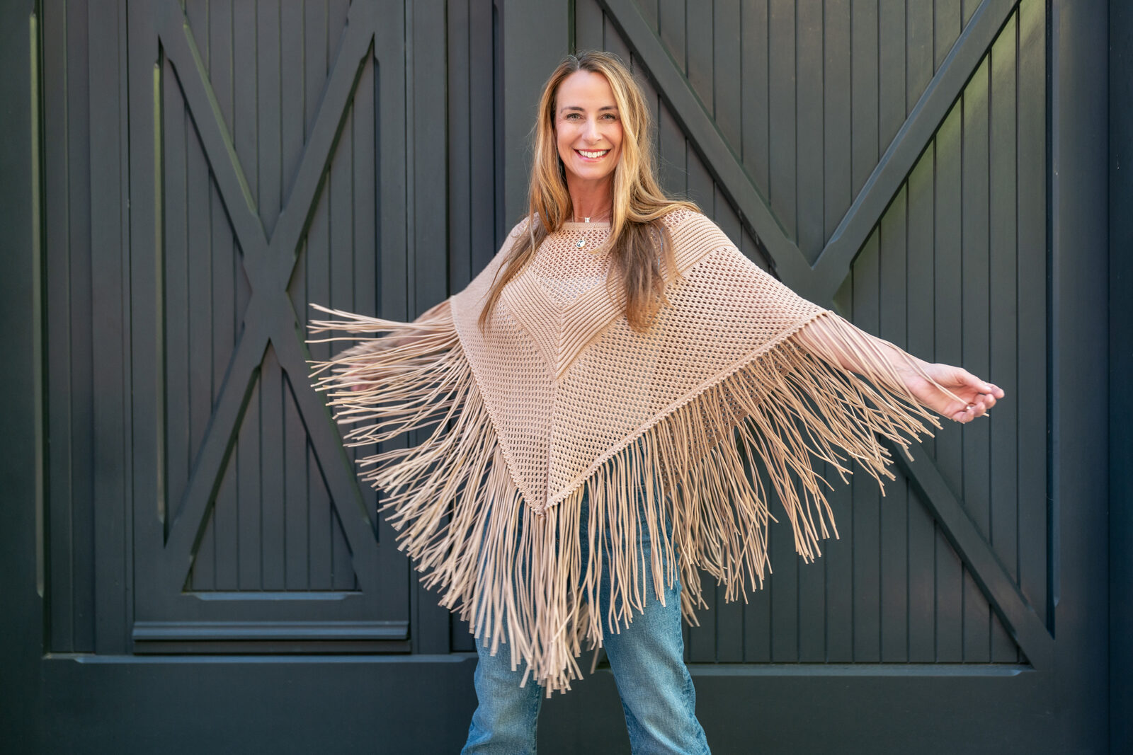 Tracey Abbott at a dark barn door, arms wide open in a cream fringe poncho, smiling