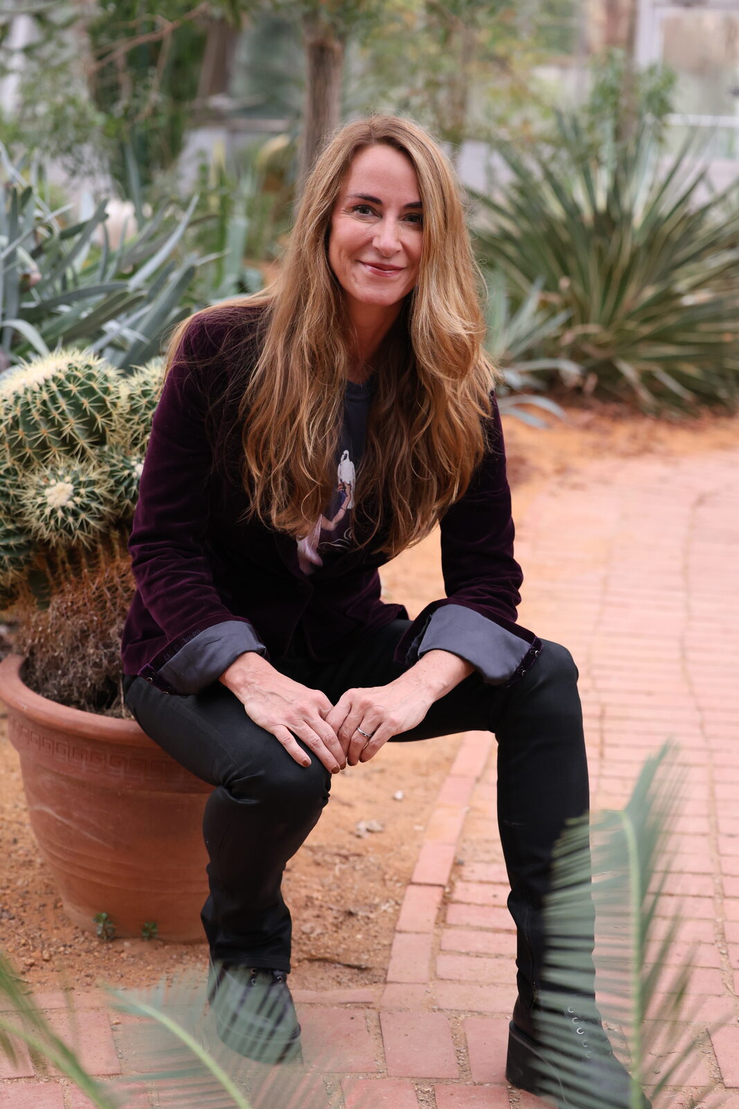 Tracey Abbott seated on a terra cotta planter among cacti, wearing a velvet blazer